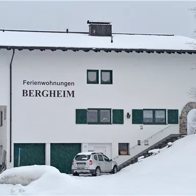 Ein großes, zweigeschossiges Gebäude mit der Aufschrift "Ferienwohnungen BERGHEIM". Die Umgebung ist schneebedeckt und winterlich.