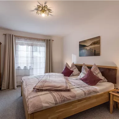 Ein gemütliches Schlafzimmer mit einem Doppelbett und dekorativen Kissen. Es gibt einen Fernseher und ein Fenster mit Vorhängen, das natürliches Licht hereinlässt.