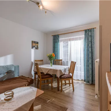 Ein gemütlicher Wohnraum mit einem Tisch und Stühlen, umgeben von natürlichem Licht. Blumenschmuck sorgt für eine einladende Atmosphäre.