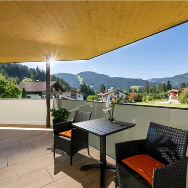 A sunny terrace with a table and two chairs. In the background, green hills and a clear blue sky can be seen.
