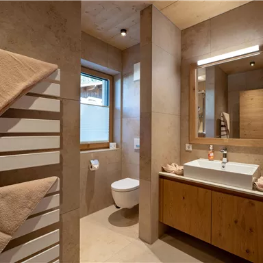 A modern bathroom with wood and stone accents. It features a sink, a toilet, and large windows for plenty of light.