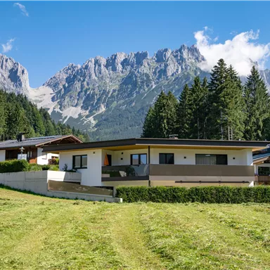 A modern house surrounded by nature, with mountains in the background. The sky is clear and the landscape is green and expansive.