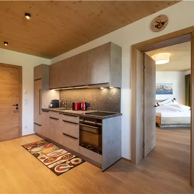 A modern kitchen with gray cabinets and high-quality appliances. In the background, a cozy living area is visible.