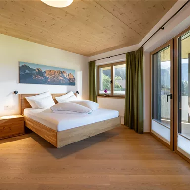 A bright bedroom with a comfortable bed and wooden decor. Large windows offer a view of nature.