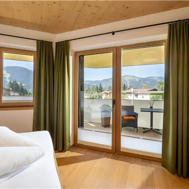 A cozy room with large windows and a view of the mountains. The space is bright and has green curtains as well as a balcony.