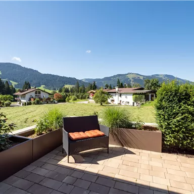 A quiet balcony with a view of a green meadow and mountains in the background. A cozy seating area with a cushion invites you to relax.