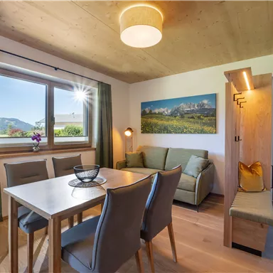 A cozy living area with a table and chairs, a sofa, and plenty of natural light. The view from the window reveals a beautiful landscape with mountains in the background.