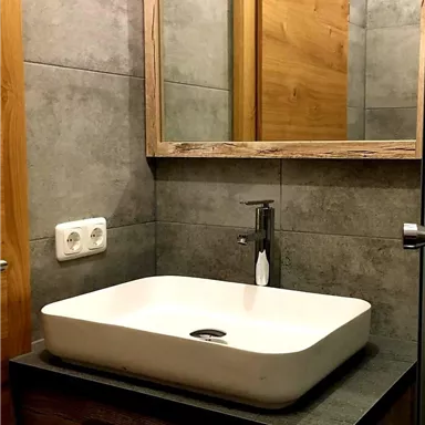 A modern bathroom with an elegant white ceramic bowl and a wooden vanity. There is a mirror on the wall, and the walls are clad with gray tiles.
