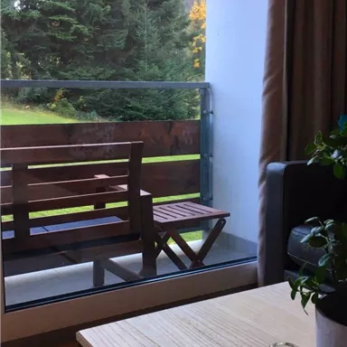 A cozy living room with a view of a balcony and green trees. The table is made of wood and a plant is placed on it.