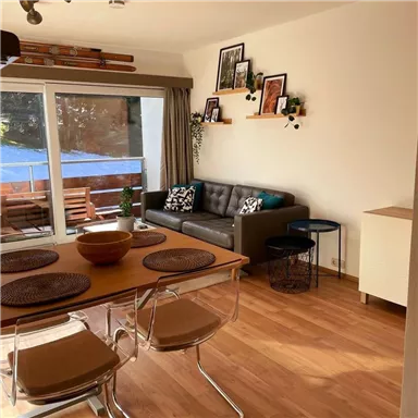 A modern furnished living room with a cozy couch and a dining table. Large windows offer a view of a terrace with garden furniture.