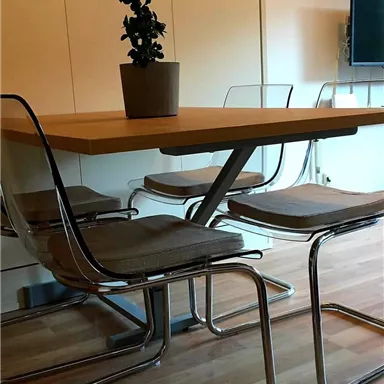 A modern dining table with four transparent chairs and a potted plant. In the background, a television is visible.