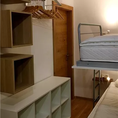 A simple room with bunk beds, shelves, and a clothing rack. The floor is wooden and the walls are painted light.