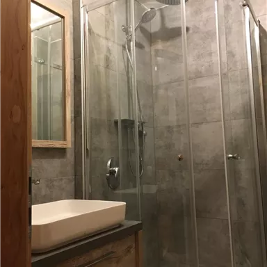 A modern bathroom with a glass shower cabin and an elegant sink arrangement. The walls are designed with gray tiles, and a wooden cabinet provides storage space.
