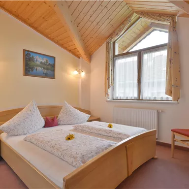 A cozy bedroom with a wooden ceiling and a large window. The bed is decorated with pillows and flowers.