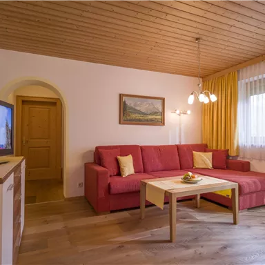 A cozy living room with a red sofa and a wooden table. Large windows let in a lot of daylight.