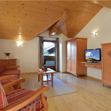 A cozy living space with a wooden ceiling and large windows. It includes a television, a sofa, and a wooden piece of furniture.