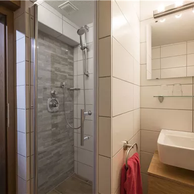A modern bathroom with a shower and wash basin. There are bright tiles and a comfortable chair in the corner.