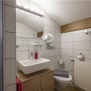 A modern bathroom with white tiles and wooden accents. It features a sink, a shower, and towels in neutral colors.