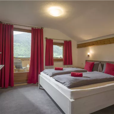 A cozy bedroom with two beds and red pillows. Large windows offer a view of the mountains and a balcony.