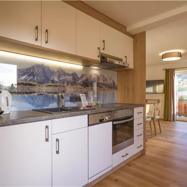 A modern kitchen with white cabinets and wood details. In the background, there is a picturesque mountain landscape.