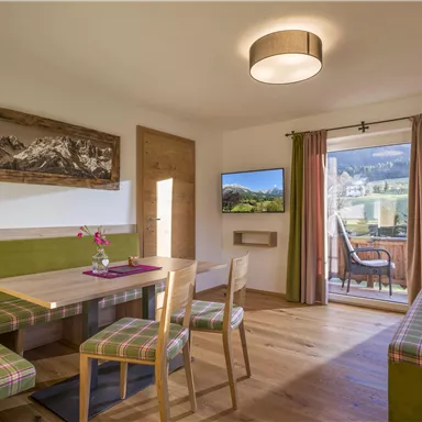A cozy living area with a wooden table, colorful seating, and a television. Large windows provide a view of the landscape.