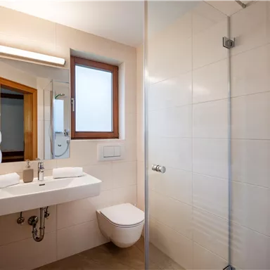 A modern bathroom with a shower, sink, and mirror. Bright tiles and natural lighting through a window create a friendly atmosphere.