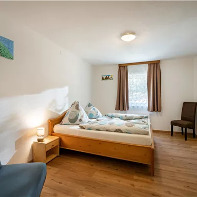 A cozy room with two single beds and a comfortable armchair. The room has bright walls, a wooden floor, and a quiet atmosphere.
