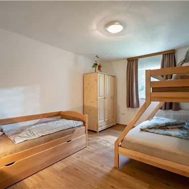 A cozy bedroom with a bunk bed and a single bed. The walls are light and the room has hardwood floors as well as a windowsill with curtains.