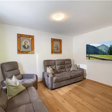 A cozy living room with two gray armchairs and a modern sofa. Nostalgic pictures hang on the wall and a television displays a beautiful landscape.