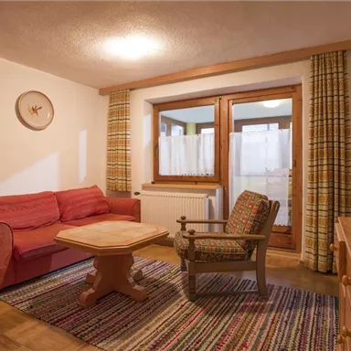 Ein gemütliches Wohnzimmer mit einem roten Sofa, einem Holztisch und einem Sessel. Die großen Fenster lassen viel Tageslicht herein und der Teppich sorgt für eine warme Atmosphäre.