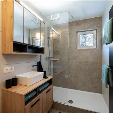 Ein modernes Badezimmer mit einer Dusche aus Glas und Holzregalen. Die Einrichtung ist schlicht und elegant, mit einem großen Spiegel und einem Waschbecken auf einem Holzschrank.
