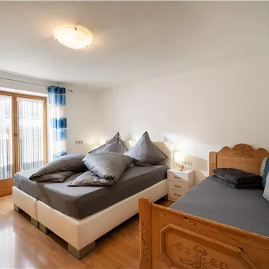 Ein helles Schlafzimmer mit einem Doppelbett und einem Einzelbett. Große Fenster lassen viel Licht herein und die Wände sind in neutralen Farben gehalten.