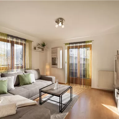 Ein modernes Wohnzimmer mit einem grauen Sofa und einem Couchtisch. Große Fenster sorgen für viel Tageslicht und eine einladende Atmosphäre.