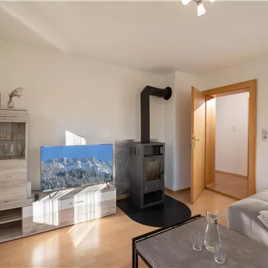 Ein modernes Wohnzimmer mit einem Holzofen und einem großen Fernseher. Helle Wände und natürliche Holzfarben schaffen eine einladende Atmosphäre.
