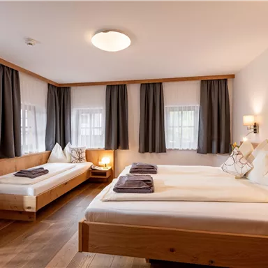 Ein gemütliches Zimmer mit zwei Betten, einem zusätzlichen Bett und Holzdetails. Die Fenster sind mit Vorhängen verhängt und es gibt warmes Licht von einer Lampe.