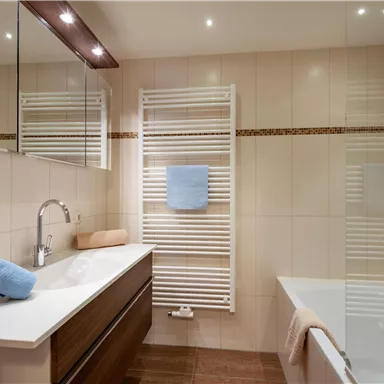 Ein modernes Badezimmer mit einer Badewanne und eleganten Holzschränken. Handtücher in sanften Farben liegen bereit, und große Spiegel reflektieren das Licht.