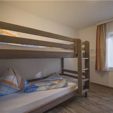Ein einfaches Zimmer mit einem Etagenbett aus Holz. Es gibt große Fenster mit Vorhängen und eine helle, freundliche Atmosphäre.