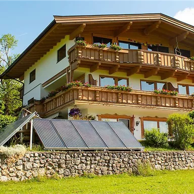 A cozy house in alpine style with a large balcony. Surrounded by green spaces and a sunny sky.