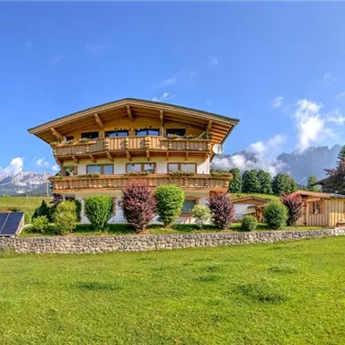 A beautiful wooden house in the Alps with large windows. The surroundings are green and planted with trees and shrubs.