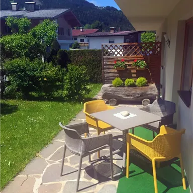 A cozy outdoor area with a table and colorful chairs on a well-maintained garden path. In the background, plants and a wooden wall can be seen, surrounded by a beautiful mountain landscape.