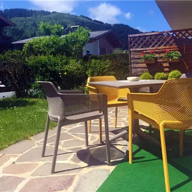 A cozy outdoor area with a table and colorful chairs. In the background, there are green bushes and a beautiful view of the mountains.