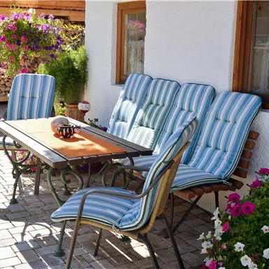 A cozy outdoor area with a table and comfortable chairs. Colorful flowers adorn the space, while a stack of wood is visible in the background.