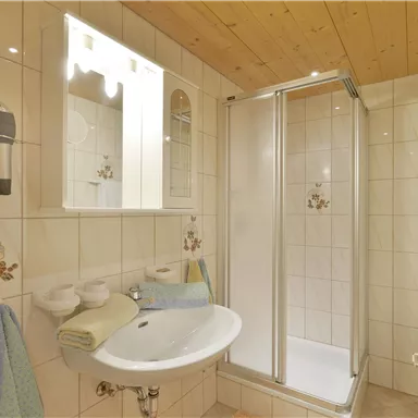 A modern bathroom with a shower cabin and sink. The walls are tiled in light colors, and there is a mirror as well as towels.