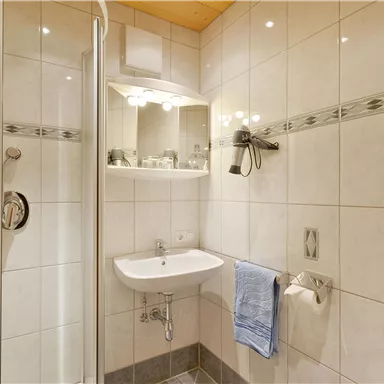 A modern bathroom with a shower, a sink, and a large mirror. The walls are brightly tiled, and blue towels are hanging.