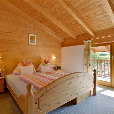 A cozy room with a wooden ceiling and a large window. The bed is decorated with colorful pillows and offers a nice view outside.