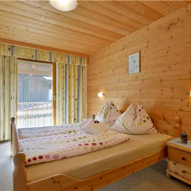 A cozy bedroom with wood paneling and a large window. There are two beds with colorful cushions and a small bedside lamp.