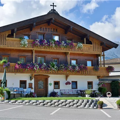 A traditional chalet with blooming balconies and wooden decorations. The surroundings are surrounded by mountains and a clear sky.