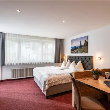 A cozy hotel room with a large bed and a table. The walls are decorated with pictures of landscapes, and there is plenty of natural light.