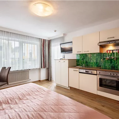 A modern room with a double bed and a kitchenette. Large windows ensure plenty of daylight.