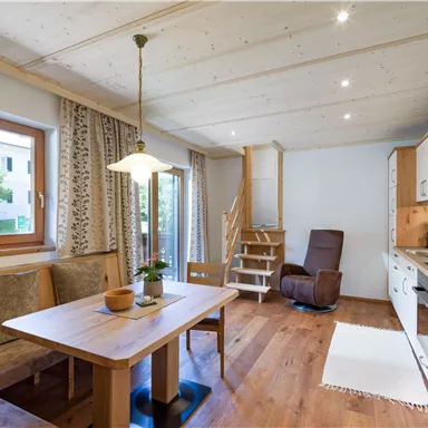 Ein gemütlicher Wohnraum mit Holztisch und Stühlen. Die Küche ist modern und hell, mit großen Fenstern und einem Treppenaufgang.